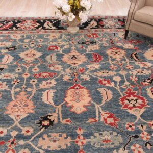 Blue traditional rug with red and blush accents lays near a light upholstered chair and flower vase.