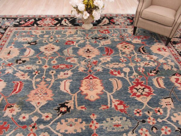 Blue traditional rug with red and blush accents lays near a light upholstered chair and flower vase.