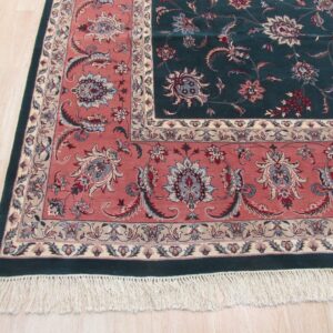 Ornate dark green and rose floral rug with cream fringe rests on light beige wood flooring.
