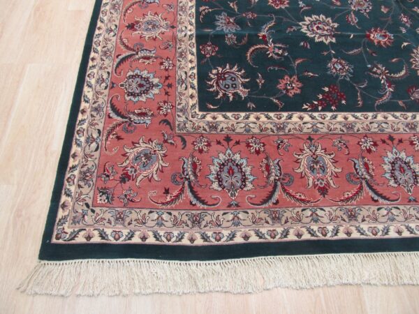 Ornate dark green and rose floral rug with cream fringe rests on light beige wood flooring.