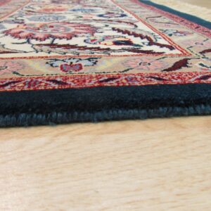 Detailed view of a low-angle rug edge showing dark blue pile, floral pattern, and light wood floor.