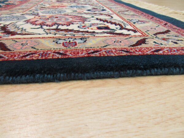 Detailed view of a low-angle rug edge showing dark blue pile, floral pattern, and light wood floor.