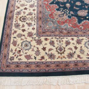 A detailed traditional rug with a teal and rust field, cream floral border, and fringes on light wood flooring.