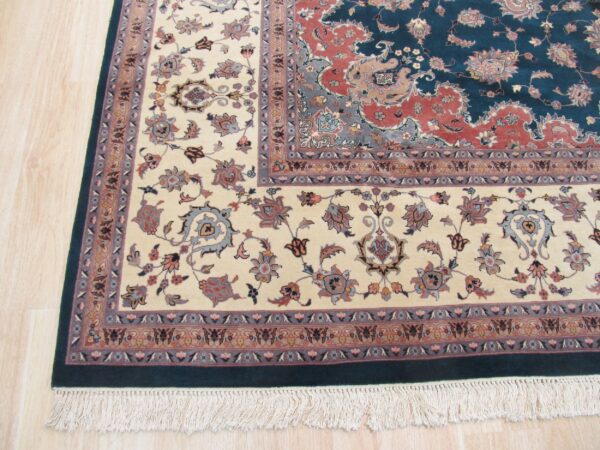 A detailed traditional rug with a teal and rust field, cream floral border, and fringes on light wood flooring.