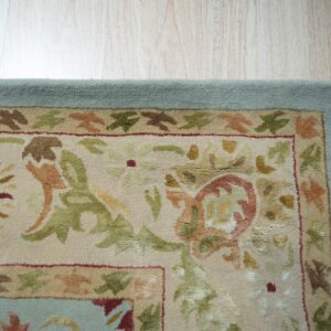 Textured rug with sage, rust, and beige floral patterns, bordered in light blue on wood planks.