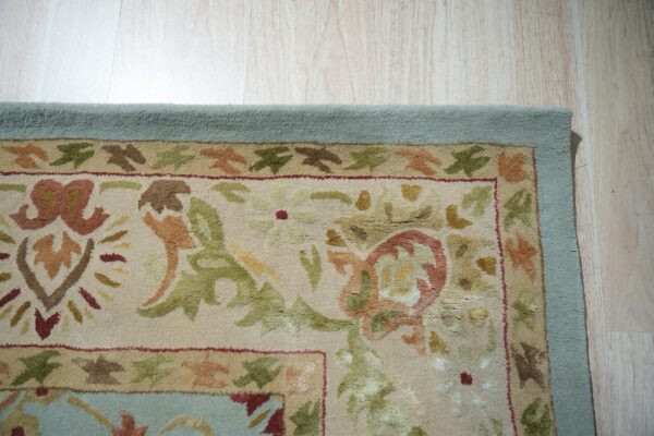 Textured rug with sage, rust, and beige floral patterns, bordered in light blue on wood planks.