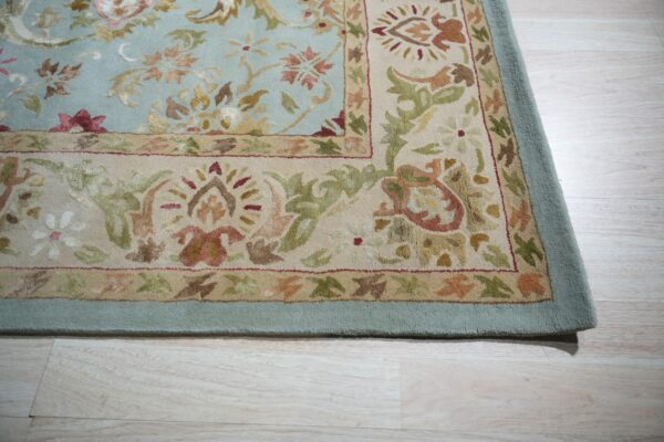 Pale blue rug with a beige and floral border lies on light blonde wood flooring.