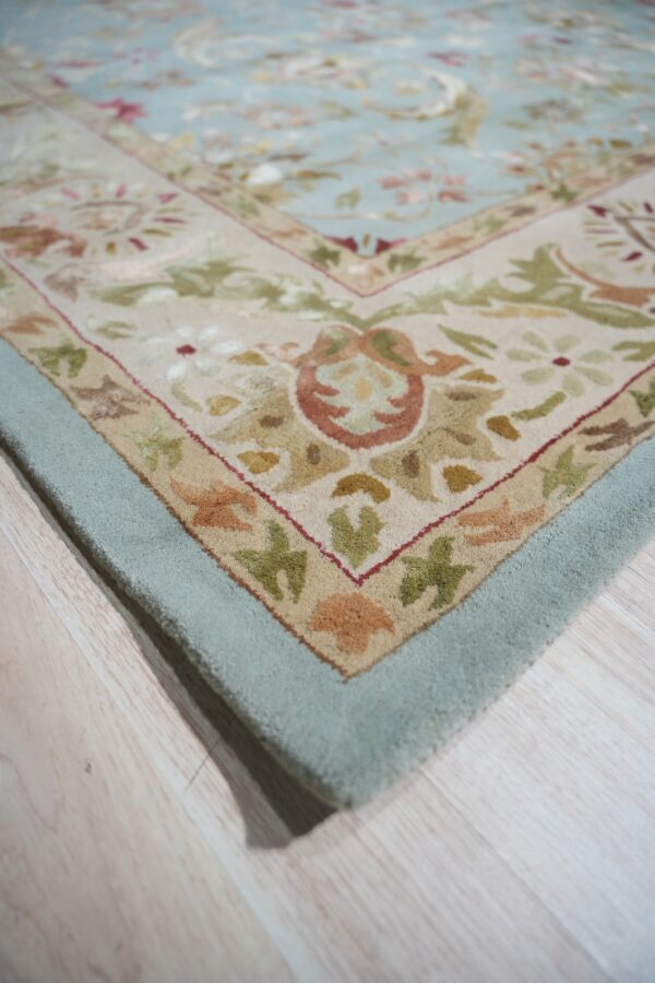 A close-up corner of a light blue floral rug with beige and rust borders on pale wood flooring.