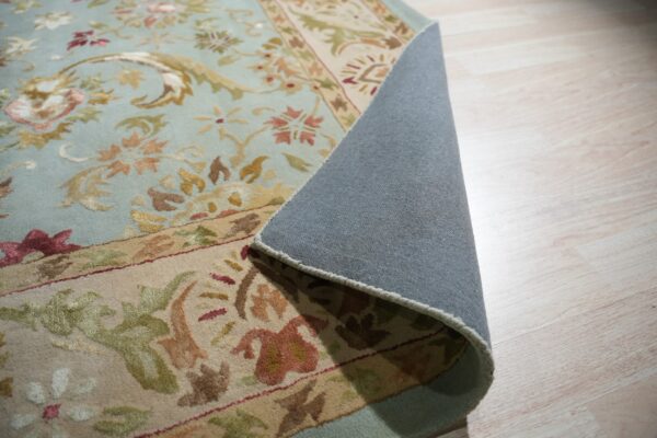 Light blue floral rug with a beige border rolled back showing gray backing on light wood floor.