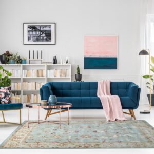 Light blue floral rug anchors a modern room with teal velvet furniture, pink curtains, and white bookshelves.