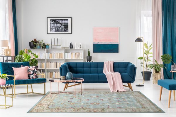 Light blue floral rug anchors a modern room with teal velvet furniture, pink curtains, and white bookshelves.