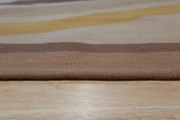 Low-angle product view of a striped flatweave rug in brown, yellow, and beige tones on light wood floor.
