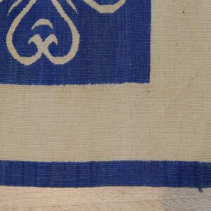 Blue flatwoven rug corner with natural beige scrolling patterns and striped borders on light wood flooring.