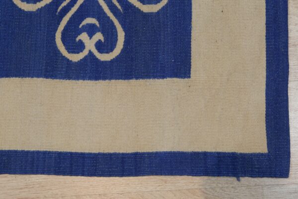 Blue flatwoven rug corner with natural beige scrolling patterns and striped borders on light wood flooring.