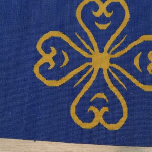 Blue flatweave rug featuring a large gold stylized clover or heart motif on light wood floor.