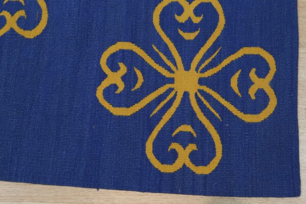 Blue flatweave rug featuring a large gold stylized clover or heart motif on light wood floor.