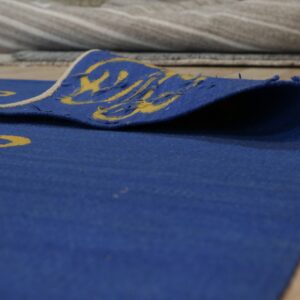 A blue rug with a yellow pattern is folded on wood flooring, showing the woven reverse texture and light edge binding.