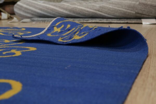 A blue rug with a yellow pattern is folded on wood flooring, showing the woven reverse texture and light edge binding.