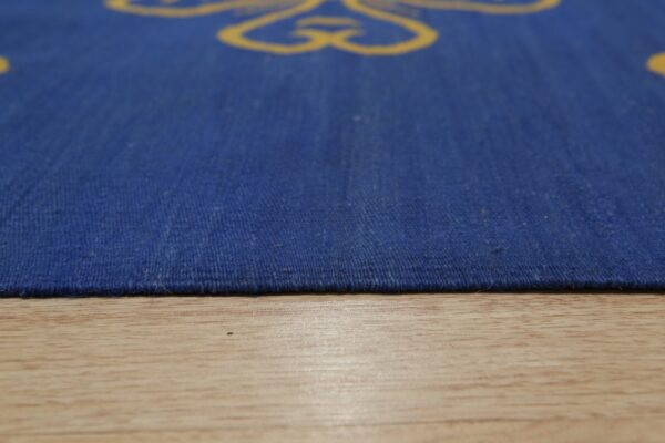 Low-angle view of a coarse, low-pile blue rug with yellow patterns resting on light wood flooring.