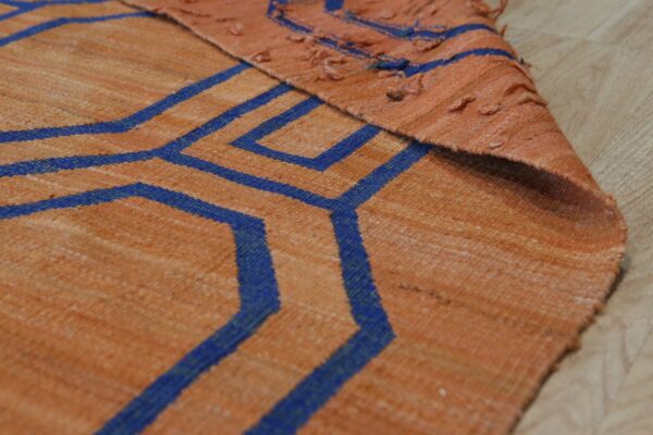 Detail shot of a flat-woven orange rug with bold blue geometric lines, folded over on wood flooring.