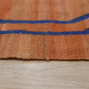 Flatweave rug detail in orange and blue geometric pattern resting on light wood flooring.