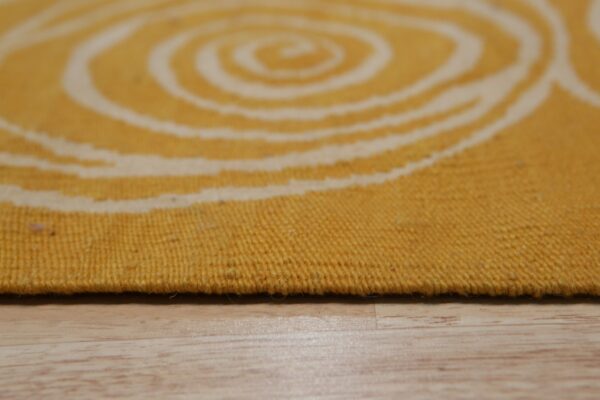 A flat-woven yellow and cream rug featuring a blurry spiral pattern rests on light wood.