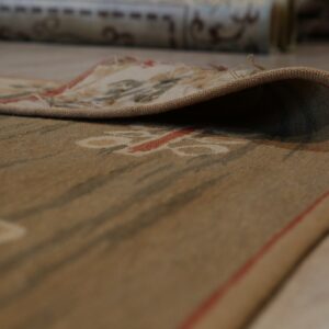 The curled edge of a low-pile tan rug with red and green botanical patterns rests on light wood flooring.