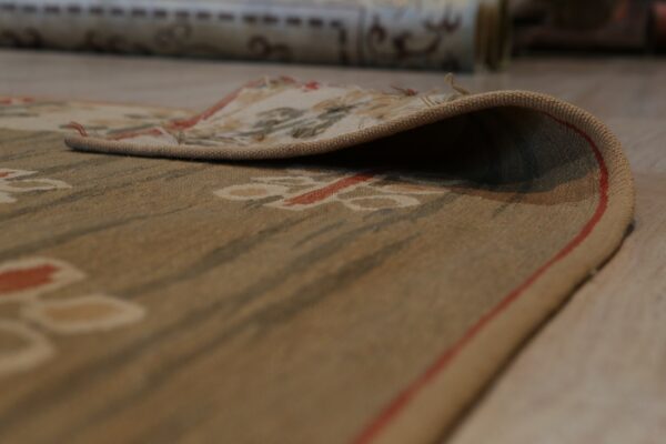 The curled edge of a low-pile tan rug with red and green botanical patterns rests on light wood flooring.