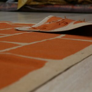 A low-pile orange and cream rug with a brick pattern is curled up on light wood flooring.