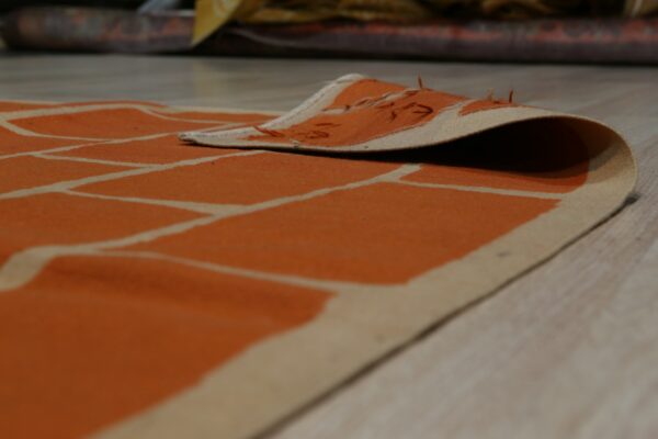 A low-pile orange and cream rug with a brick pattern is curled up on light wood flooring.
