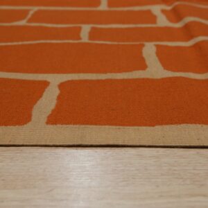 Orange and tan flatweave rug featuring an abstract brick pattern rests on light wood planks.