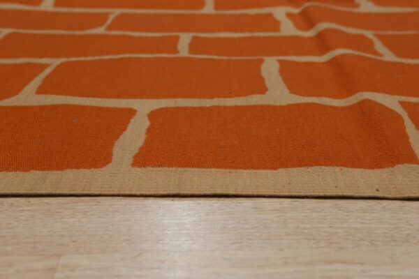 Orange and tan flatweave rug featuring an abstract brick pattern rests on light wood planks.