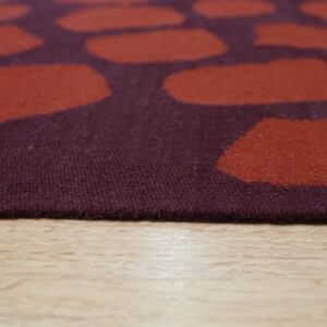 Close-up of a low-pile rug featuring large rust shapes on a dark plum background over light wood flooring.