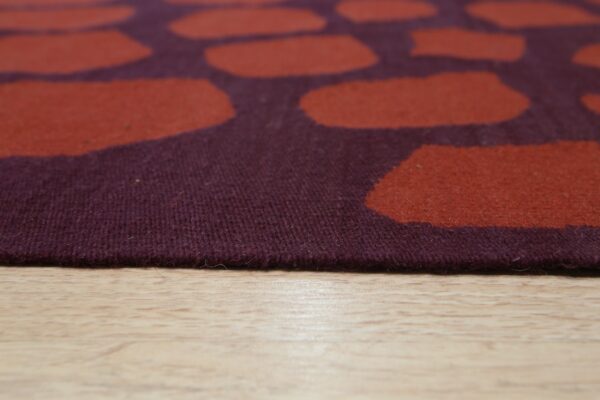 Close-up of a low-pile rug featuring large rust shapes on a dark plum background over light wood flooring.