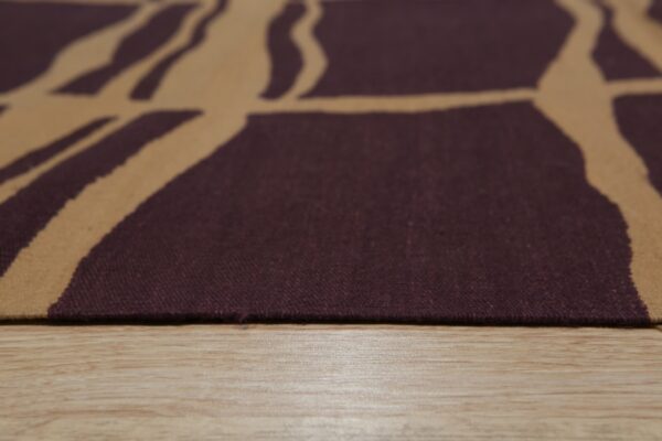 Flatweave rug with a maroon and tan abstract pattern rests closely on light wood flooring.
