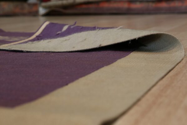 Close-up showing the curled edge of a purple and beige rug on a light wood floor.