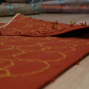 Close-up of a low-pile rust rug featuring yellow outlines, folded over light wood flooring.