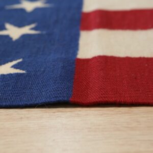 Close-up of a blue, red, and cream flat-weave rug with stars and stripes resting on a light wood surface.