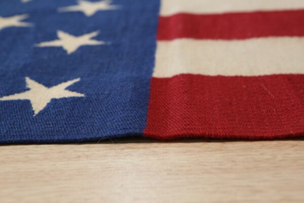 Close-up of a blue, red, and cream flat-weave rug with stars and stripes resting on a light wood surface.