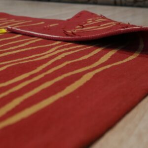 Red flatweave rug with gold wavy stripes folded over on light wood flooring.