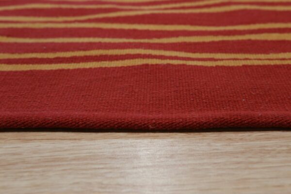 Red flatweave rug with wavy gold stripes rests closely on light wooden plank flooring.