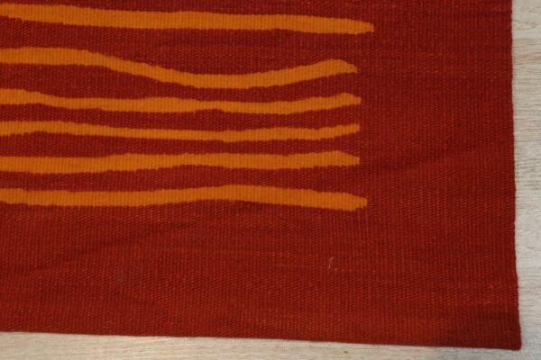 A rust-colored flatweave rug with abstract orange horizontal stripes rests on light wood flooring.