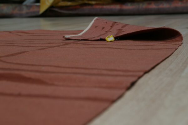 Terracotta low-pile rug with subtle stripes, partially folded back, showing a yellow tag on light wood flooring.