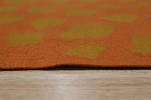 Orange flatweave rug with abstract yellow spots rests on a light wood grain surface.