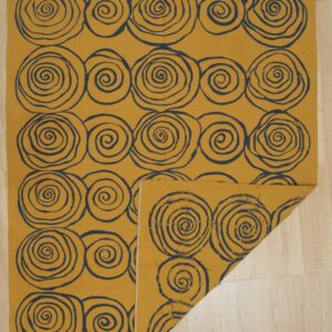 Golden yellow flatweave rug with large dark blue spiral patterns is partially folded on pale wood flooring.