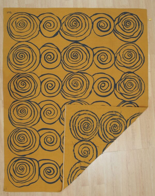 Golden yellow flatweave rug with large dark blue spiral patterns is partially folded on pale wood flooring.