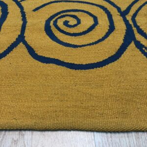 Mustard flatweave rug with thick navy spiral patterns resting on distressed light wood planks.