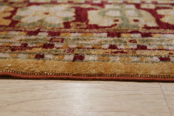 The edge of a medium-pile rug features red, gold, and green traditional patterns over light wood flooring.