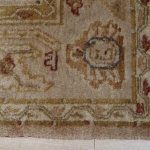 Close-up of a textured beige rug with traditional gold and maroon patterns resting on light wood plank flooring.