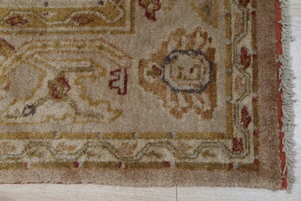 Close-up of a textured beige rug with traditional gold and maroon patterns resting on light wood plank flooring.